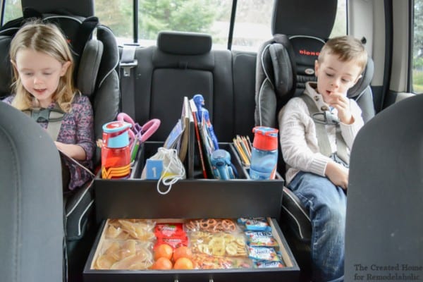 Two Kids In Car Seats With Books, Sippy Cups, And Snacks In An Organizer Between Them