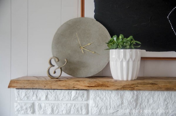 Simple DIY Concrete Clock With Gold Hands Sitting On Wooden Shelf Above White Brick Mantle And White Planter With Greenery
